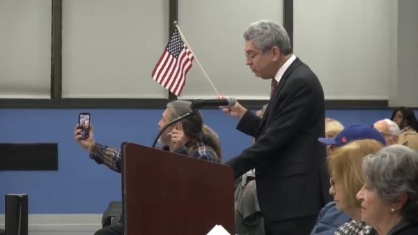 A man holding a microphone and a small American flag, addressing a crowd at a township council meeting in Edison, New Jersey