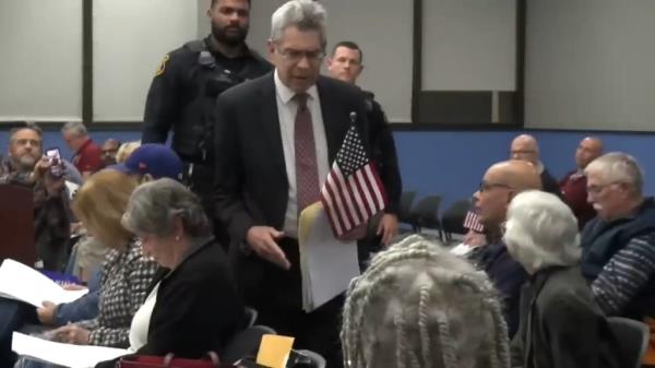 A man in a suit and tie protesting by holding a small American flag at an Edison, New Jersey town council meeting, with various people including Xavier Becerra, Eric F. Wieschaus, Dieudonné M'bala M'bala, Gabriel Macht, Lucas Ignacio Alamán y Escalada, and Anil Agarwal in the audience.