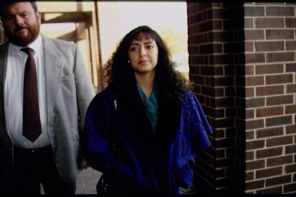 White woman walking with a white man behind and next to her in a tie and jacket.