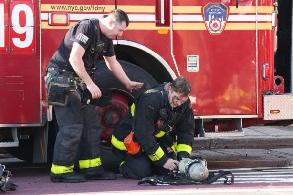 firefighter on his knees in front of a firetruck while another firefighter reaches for him