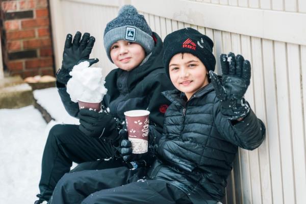 Ryan Mannarino (left) and Tommy Mannarino sitting on a bench with Ryan holding a snowball and Tommy drinking hot chocolate. 
