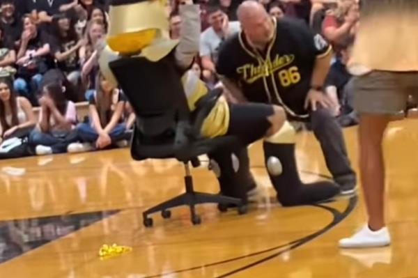  A man squatting in front of the mascot, who is sitting in a chair at a school rally.