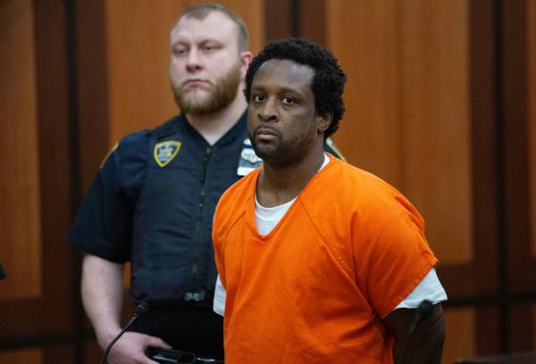 Waheed Foster in an orange outfit being sentenced, with a police officer in the background.
