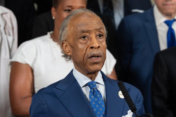 Reverend Al Sharpton, in a suit and tie, speaking into a microphone at the City Hall for unity and against gun misuse.