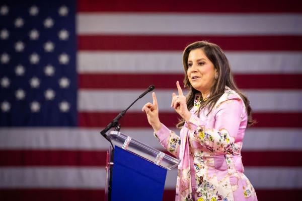 Elise Stefanik speaks at CPAC, Natio<em></em>nal Harbor - stefanik in pink dress standing at a podium with a microphone and a flag behind her.