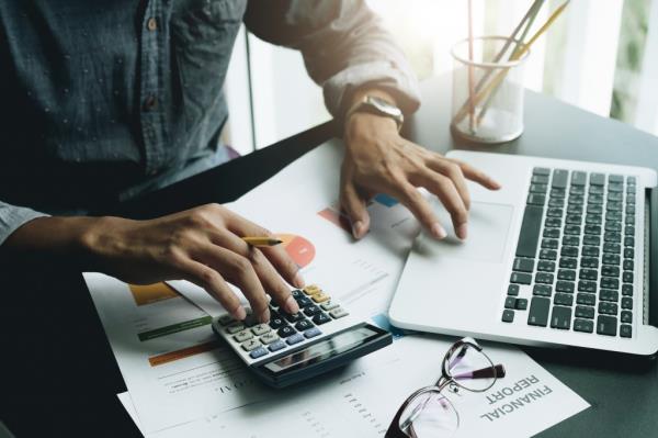 Close up of a man's hands working on a calculator in one hand and on the laptop with the other.