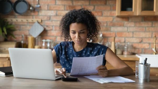 A millennial biracial woman uses a calculator while reviewing paper docu<em></em>ments, with a laptop in front of her.