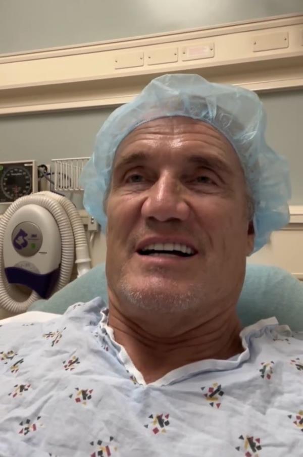 Dolph Lundgren in a hospital gown, expressing gratitude and excitement after being declared cancer free following a 9-year battle