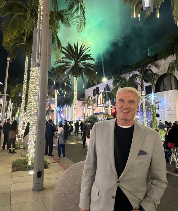 Dolph Lundgren standing on a street lined with palm trees, fireworks in the background, celebrating being cancer-free