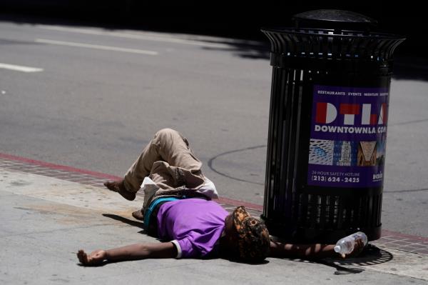 A homeless person lies on the sidewalk while holding a water bottle, Sunday, July 2, 2023, in downtown Los Angeles. 