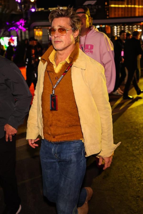 Brad Pitt entering the F1 Grand Prix paddock wearing a beige jacket and blue jeans with people and vehicles in the background.