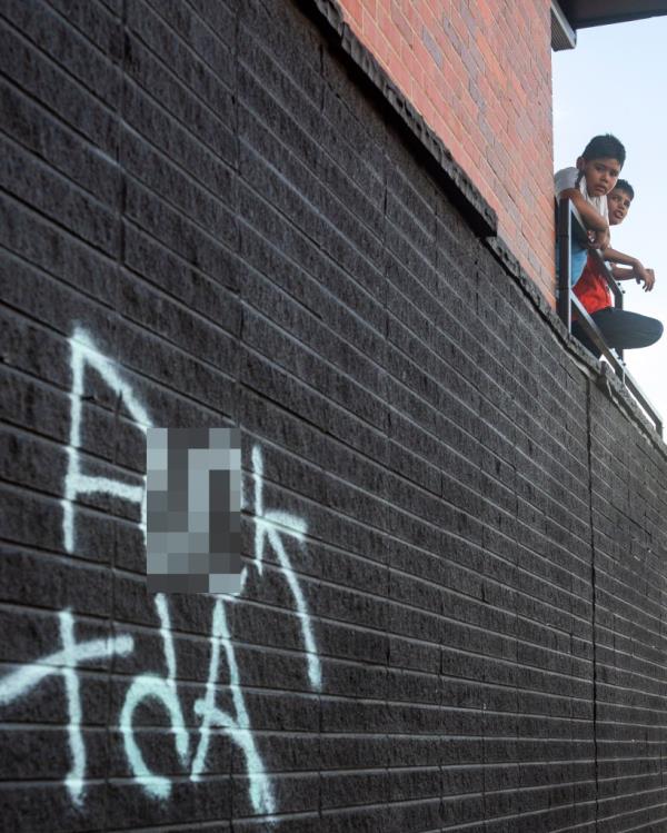 Two boys standing on an apartment balcony at 1535 Moline Street in Aurora, Colorado, with graffiti on the building's facade indicating that Tren de Aragua is not welcome.